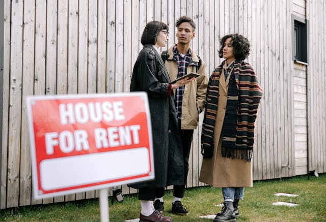 Landlord speaking with tenants on property behind a "House For Rent" sign